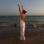 Woman in a sparkly dress standing on a beach with arms raised at sunset.