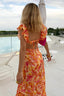 Woman in a colorful dress standing by a poolside with chairs and umbrellas in the background.